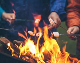 A group of people roast marshmallows over a fire pit. They practice good fall fire safety by keeping a safe distance.